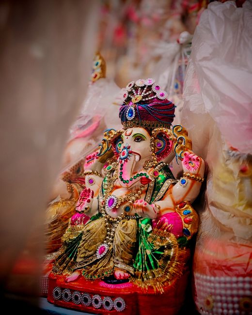 A colorful, intricately decorated statue of Lord Ganesha, adorned with vibrant jewelry, a multicolored turban, and floral garlands, seated on a red base surrounded by soft pink and gold fabrics. The s