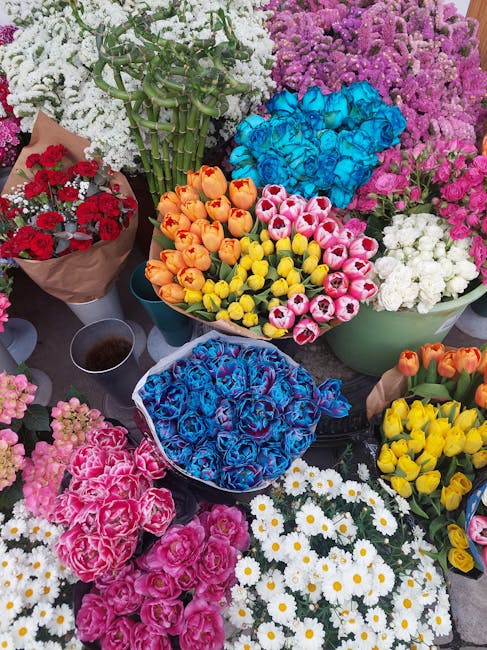 A colorful display of fresh flowers arranged in various bouquets and containers at a flower stall. The arrangement includes bright yellow, orange, pink, blue, white, and purple blooms such as tulips,