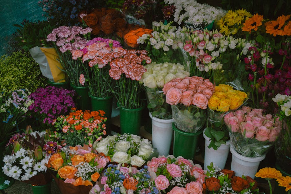 A vibrant display of fresh flowers arranged in various bouquets and buckets, featuring pink roses, white roses, orange and yellow blooms, purple chrysanthemums, white daisies, and pink carnations. The