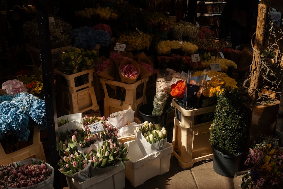 An outdoor display of fresh flowers arranged in white and black buckets and wooden crates, featuring a variety of blooms including pink tulips, blue hydrangeas, yellow chrysanthemums, and orange roses