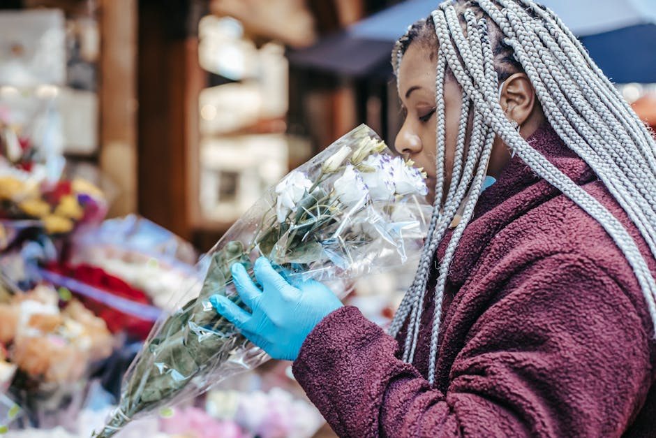 A woman with braided silver hair, wearing a maroon fleece jacket and a blue glove, holds a bouquet of white flowers wrapped in clear plastic as she appears to smell or inspect them at a flower market 