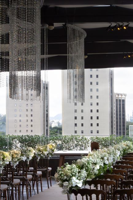 A spacious indoor venue with large floor-to-ceiling windows offering views of tall modern buildings and a cloudy sky. The room is decorated with a long rectangular table adorned with numerous white fl