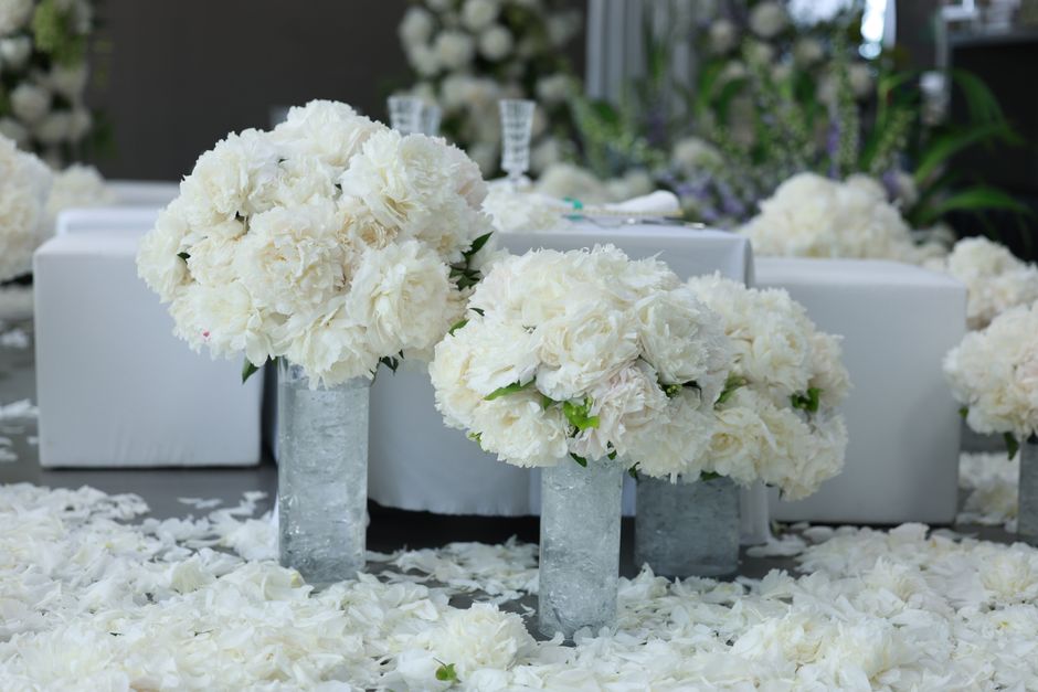 Close-up of two elegant floral arrangements featuring large, lush bouquets of white peonies and roses with delicate petals and fresh green leaves, arranged in tall, cylindrical glass vases with a text