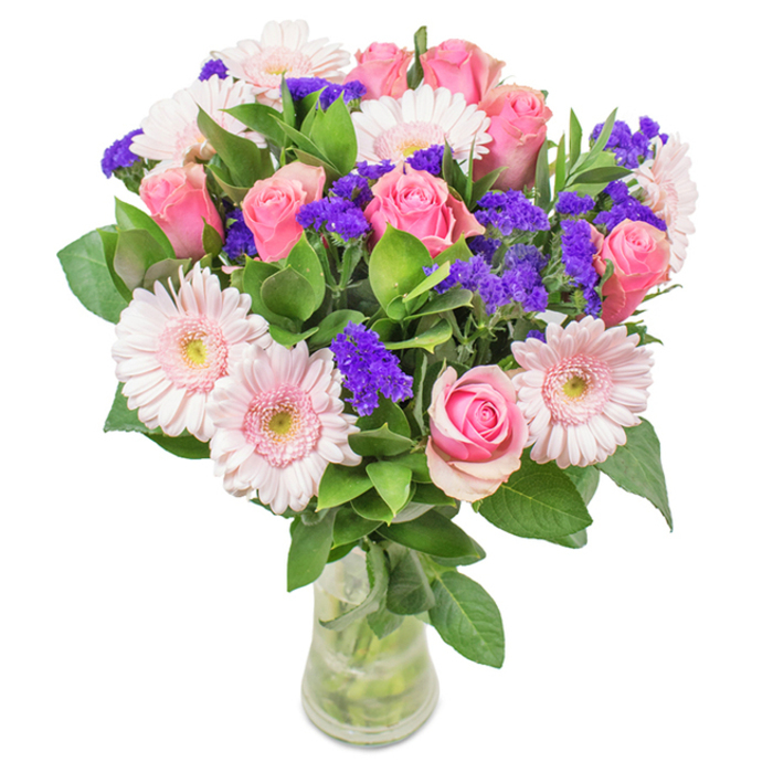 An artfully arranged bouquet, presented in a pristine clear glass vase against a simple white backdrop-the very essence of modern Elephant and Castle style. The arrangement features approximately 7-9 velvety pink roses, some with petals gently furled, recalling the classic spiral at the heart of every fine bloom. These roses are complemented by about 5-7 pale pink Gerbera daisies, distinguished by their wide-faced petals and luminous, almost whisper-white centres. Vivid purple statice clusters offer a playful texture and rich contrast, their colour reminiscent of local murals scattered near Southwark landmarks. Deep green foliage surrounds the blooms, lending a sense of abundance and natural freshness, mirroring the foliage of the nearby Pasley Park. The vase's transparent sides reveal vibrant green stems and shimmering water, heightening the sense of just-delivered freshness-a popular choice for occasions from birthdays to Mother's Day throughout the SE1 community. This bouquet merges artistry with heartfelt intent, inviting celebration, joy, and everyday beauty to the spaces and stories of Elephant and Castle.